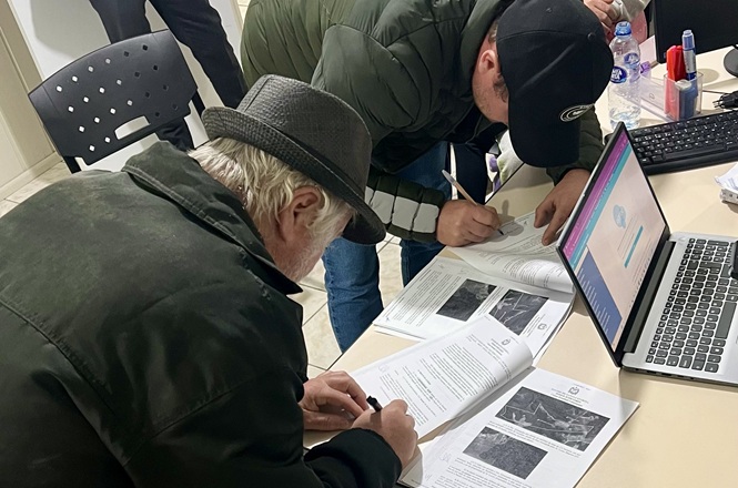 Foto horizontal: moradores assinam documentos durante atendimento de regularização fundiária, com formulários e computador sobre a mesa.