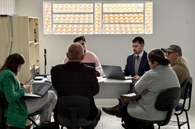 Foto horizontal: equipe técnica atende moradores em reunião de regularização fundiária, com análise de documentos e uso de computadores em ambiente comunitário.