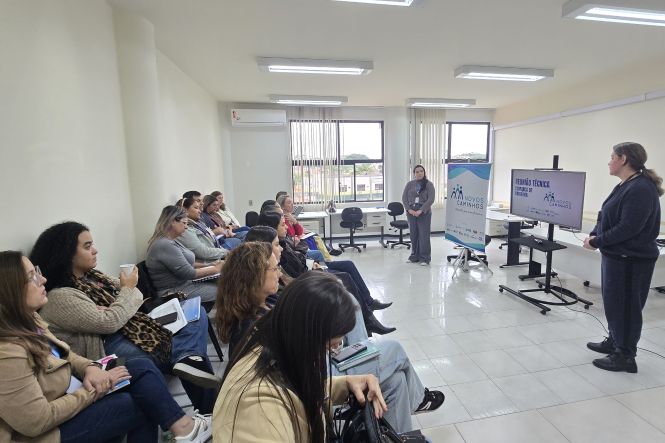 Foto horizontal: participantes acompanham apresentação do programa Novos Caminhos em sala de reunião, facilitadoras ao frente, telão ao lado, ambiente institucional, clima de aprendizagem e diálogo.