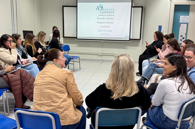 Foto horizontal: participantes em roda de conversa durante reunião do programa Novos Caminhos no Vale do Itajaí, apresentação projetada ao fundo, ambiente educativo, clima de diálogo e troca.