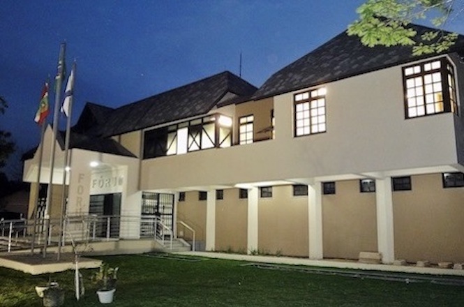 Foto horizontal: fachada iluminada do Fórum da Comarca de Pomerode, com bandeiras e céu azul-escuro ao fundo.