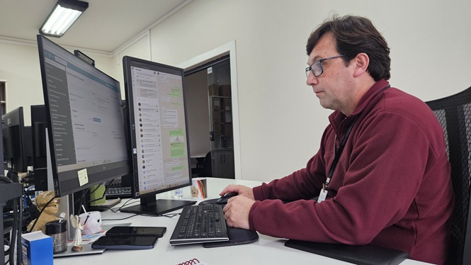 Foto horizontal: servidor trabalha diante de dois monitores de computador em ambiente de escritório.