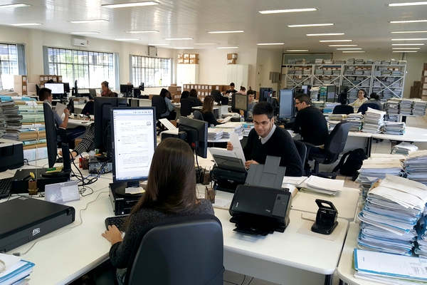 Foto horizontal: servidores trabalham em ambiente compartilhado, cercados por computadores e pilhas de processos físicos.
