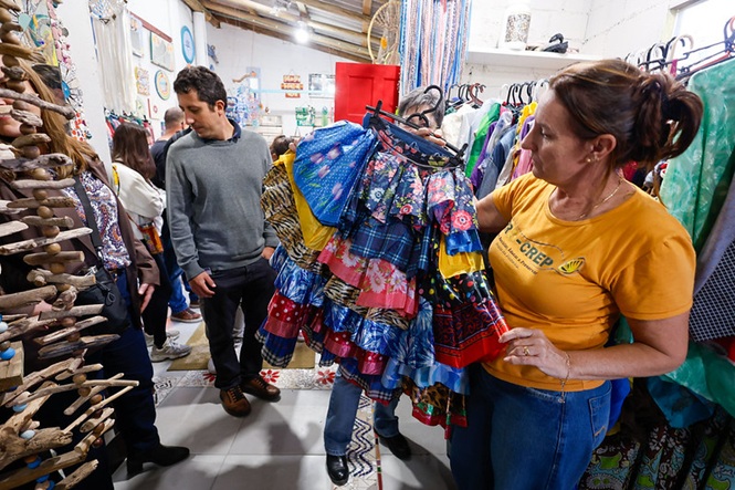 Foto horizontal: pessoas observam e selecionam roupas em espaço coletivo, peças coloridas em destaque, clima de reaproveitamento e economia solidária.