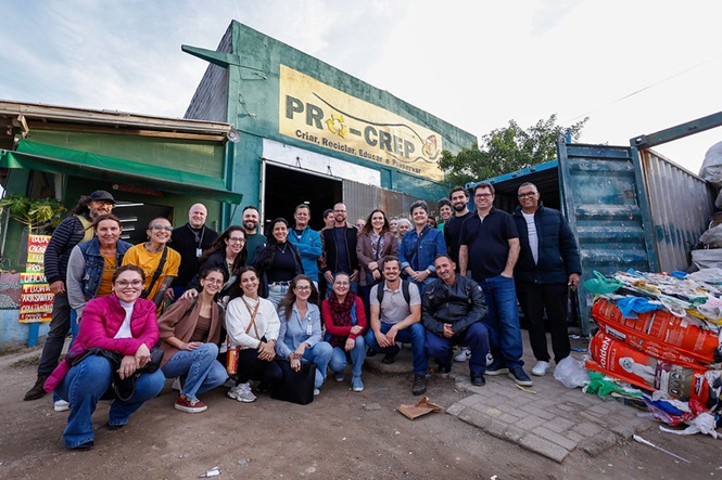 Foto horizontal: grupo posa em frente a cooperativa de reciclagem, materiais ao redor, clima de parceria, sustentabilidade e ação comunitária.