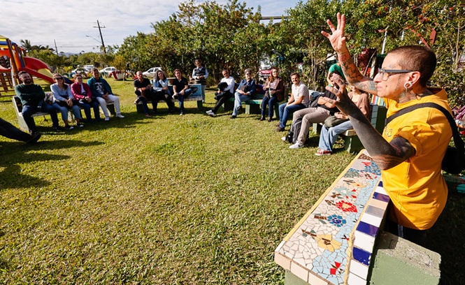Foto horizontal: grupo participa de roda de conversa ao ar livre, facilitador em destaque, clima de escuta, diálogo e construção coletiva.