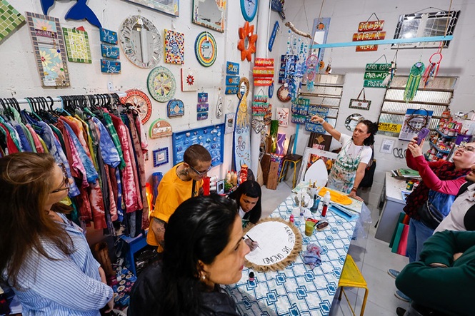 Foto horizontal: pessoas participam de atividade artesanal em espaço coletivo, objetos decorativos e reaproveitamento em destaque, clima de criatividade e economia solidária.