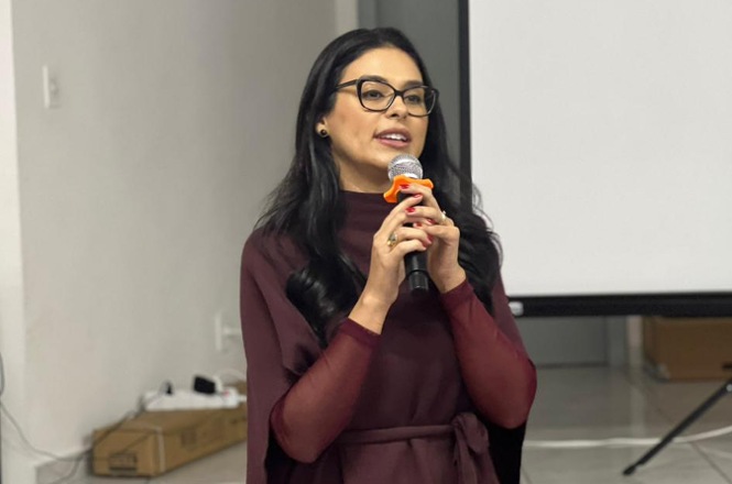 Foto horizontal: juíza Nathana Campos Dias de Souza fala ao microfone durante palestra, vestindo traje em tom bordô e óculos.