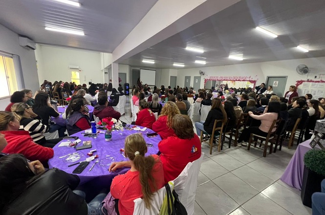 Foto horizontal: mulheres participam de encontro em salão decorado em tons de roxo e rosa, reunidas em mesas e cadeiras diante de uma apresentação.