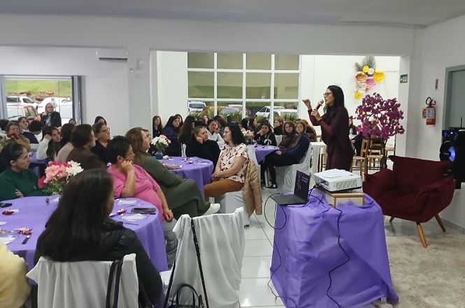 Foto horizontal: juíza Nathana Campos Dias de Souza fala ao microfone para mulheres reunidas em mesas decoradas em tons de roxo, durante palestra.