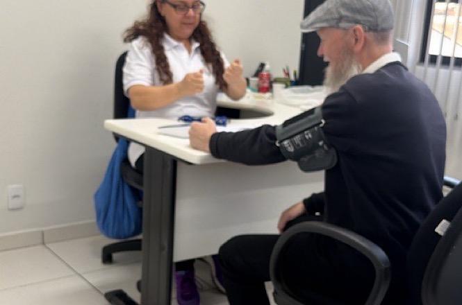 Foto horizontal: profissional de saúde em camisa polo branca orienta servidor idoso de boina cinza e roupas pretas durante aferição de pressão arterial em mesa branca de sala clara durante ação de atendimento institucional.