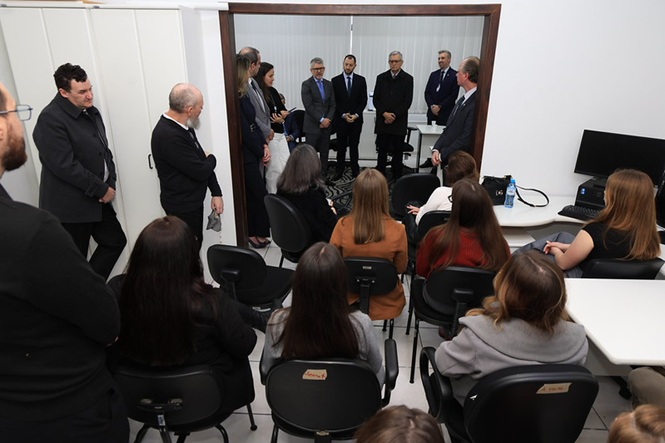 Foto horizontal: autoridades conversam com servidores reunidos em sala durante visita institucional a um setor de trabalho.