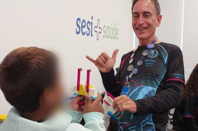 Foto horizontal: instrutor interage com aluno durante atividade de robótica educacional, protótipo em mãos, ambiente do SESI Saúde ao fundo, clima de aprendizado, incentivo e inclusão tecnológica.