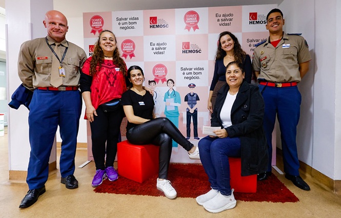 Foto horizontal: doadores e policiais militares posam no Hemosc diante de painel sobre doação de sangue.