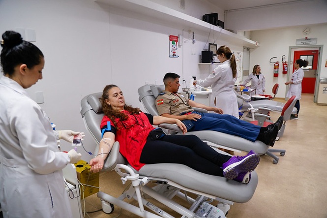 Foto horizontal: mulher e policial militar doam sangue enquanto profissionais da saúde realizam atendimento em hemocentro.