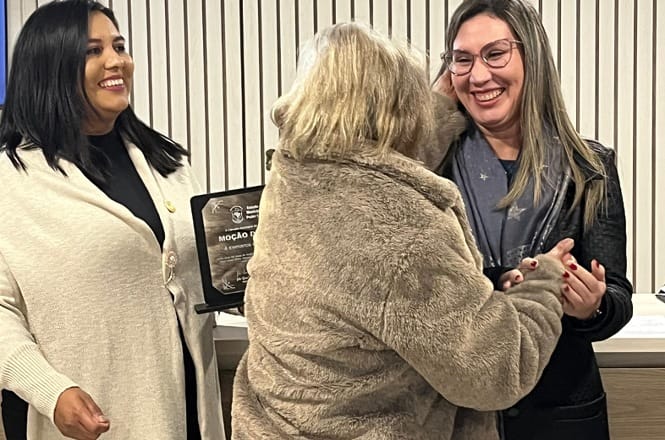 Foto horizontal: mulher homenageada recebe cumprimento de representantes do Legislativo durante cerimônia de entrega de moção de aplausos.