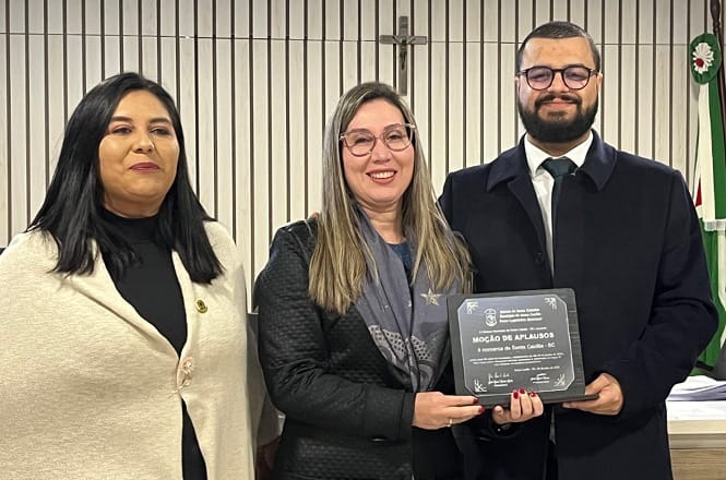 Foto horizontal: três pessoas posam durante entrega de moção de aplausos, com placa de homenagem em destaque e símbolos institucionais ao fundo.