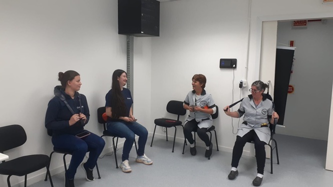 Foto horizontal: quatro mulheres participam de atividade em sala, sentadas em círculo, enquanto duas delas seguram cordas nas mãos.