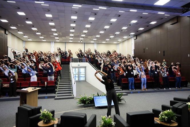 Foto colorida: participantes praticando alongamento coletivo em auditório institucional, ação de bem-estar, exercitando-se em preto, vermelho e cinza