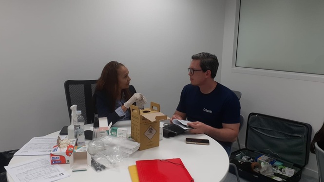 Foto horizontal: profissional de saúde conversa com homem sentado à mesa, onde estão materiais e equipamentos para atendimento do programa Saúde Itinerante.
