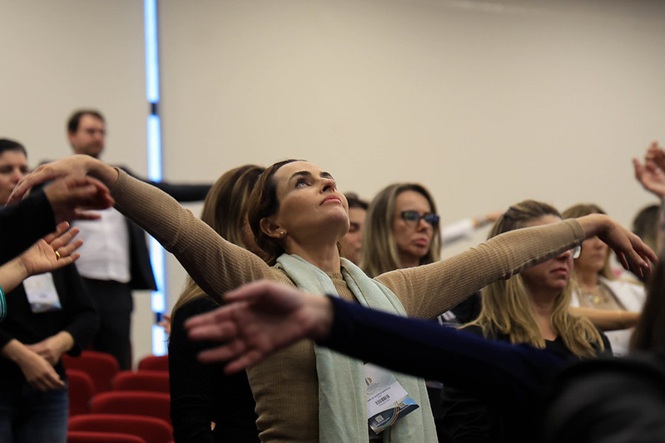Foto colorida: participantes praticando alongamento em auditório institucional, evento de bem-estar, exercitando-se em bege, vermelho e cinza