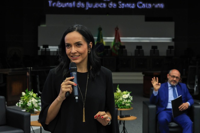 Izabella Camargo, de preto, fala ao microfone; ao fundo, André Dacol, de azul, acena plenário do TJSC, evento institucional, apresentando tema em preto, azul e branco