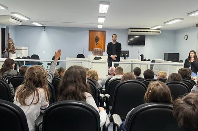 Foto horizontal: magistrado fala ao público jovem em plenário, estudante levanta a mão para participar, atividade educativa em andamento, ambiente institucional.
