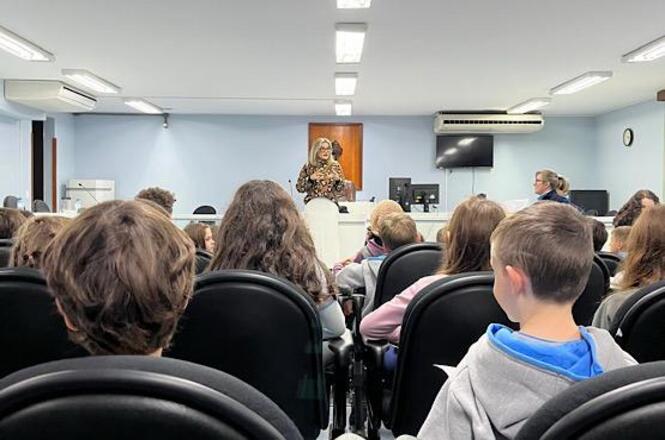 Foto horizontal: mulher fala ao público jovem em plenário, estudantes sentados acompanham atentos, atividade educativa em andamento, ambiente institucional.