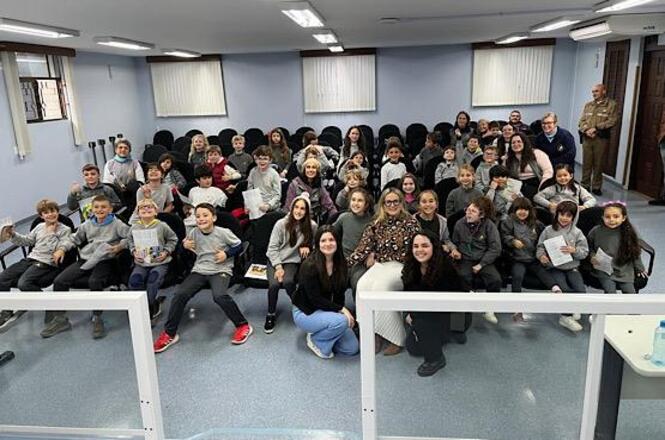 Foto horizontal: grupo de estudantes posa com equipe em plenário após atividade educativa, participantes sorridentes, ambiente institucional, clima de integração e aprendizado.