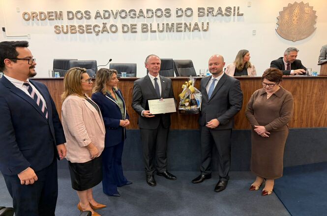 Foto horizontal: o desembargador Rubens Schulz segura placa de homenagem durante solenidade na OAB Subseção de Blumenau, ao lado de autoridades, entrega simbólica em destaque