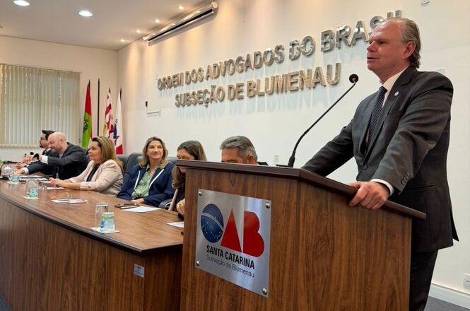 Foto horizontal: o desembargador Rubens Schulz discursa ao púlpito durante solenidade na OAB Subseção de Blumenau, autoridades ao lado acompanham a fala, ambiente institucional