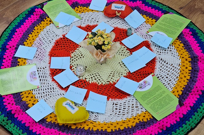 Foto horizontal: materiais dispostos em círculo sobre tapete colorido, cartões e mensagens ao redor de flores ao centro, dinâmica de grupo, clima acolhedor e reflexivo.