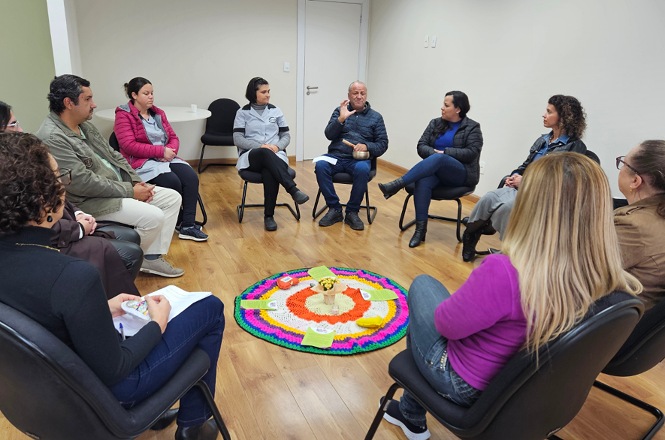 Foto horizontal: grupo de participantes em roda de conversa, homem fala ao centro enquanto demais escutam, materiais dispostos no chão, ambiente interno, clima de diálogo e reflexão.
