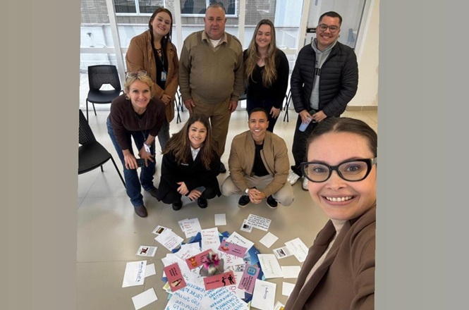 Foto horizontal: grupo posa para selfie após dinâmica em grupo, cartões espalhados no chão ao centro, ambiente interno, clima de integração e acolhimento.