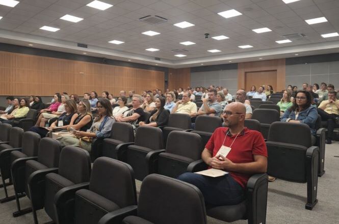 Foto colorida: público sentado em auditório amplo, seminário institucional, assistindo palestra em tons cinza e bege