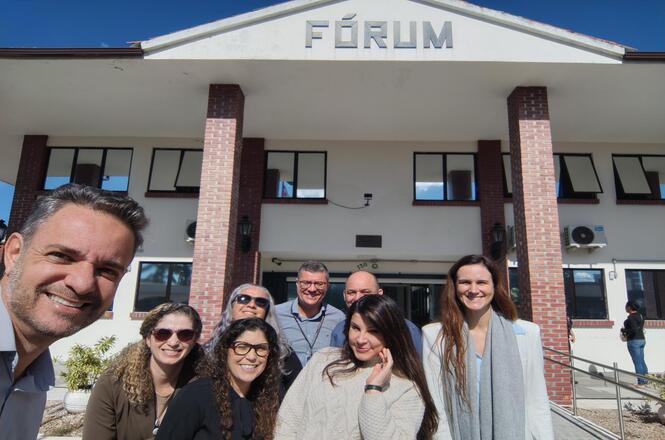 Foto horizontal: grupo registra visita em frente ao fórum, prédio institucional ao fundo, clima de integração e aproximação institucional.