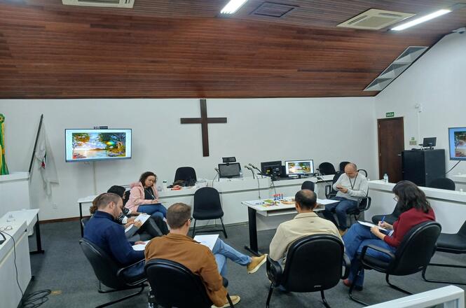 Foto horizontal: participantes reunidos em dinâmica de grupo em sala institucional, sentados em círculo, clima de escuta e construção coletiva.