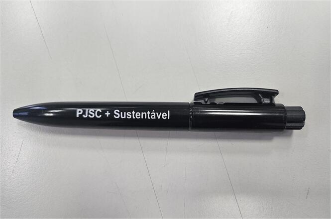 Horizontal: Caneta preta personalizada com a inscrição “PJSC + Sustentável”, posicionada sobre superfície clara. A imagem destaca o material institucional utilizado em ações de conscientização ambiental e sustentabilidade promovidas pelo Poder Judiciário de Santa Catarina.