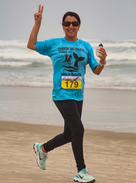 Mulher participa de corrida na praia, faz sinal de vitória e segura água, clima de superação e bem-estar