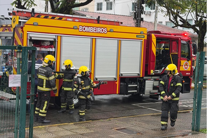 Foto horizontal: bombeiros com uniformes pretos e capacetes amarelos atuam ao lado de caminhão vermelho de resgate.