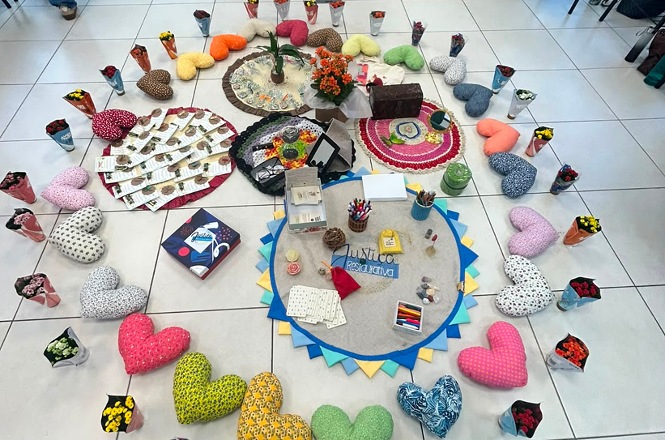 Foto horizontal: materiais lúdicos, artesanato, flores e almofadas em formato de coração organizados em círculo para dinâmica de grupo.