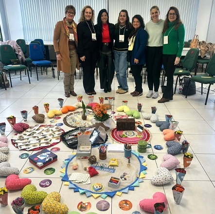 Foto horizontal: grupo posa ao redor de atividade coletiva com objetos, artesanato e materiais lúdicos utilizados em oficina de capacitação.