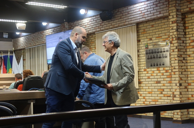 Foto horizontal: dois homens se cumprimentam com aperto de mãos; um deles segura certificado durante solenidade em plenário.