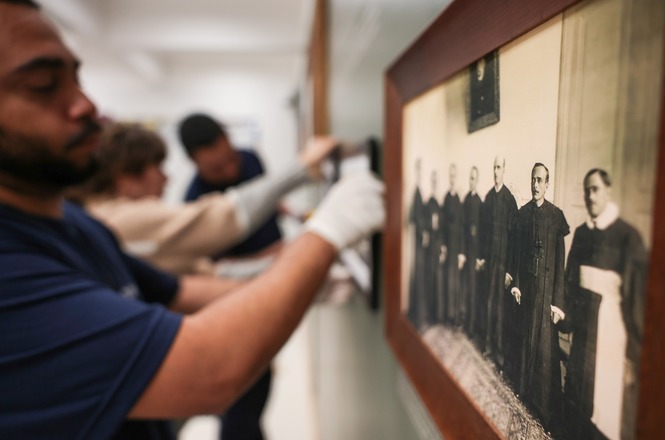 Foto horizontal: equipe com luvas instala fotografia histórica emoldurada em parede de exposição do TJSC.