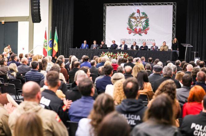 Foto horizontal: plenário da Assembleia Legislativa com autoridades à mesa e público numeroso, cerimônia oficial em andamento, ambiente institucional, clima solene.