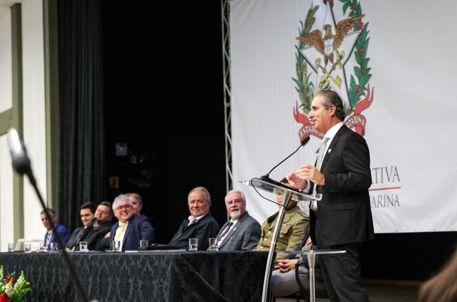 Foto horizontal: autoridade discursa em púlpito durante evento institucional, mesa de autoridades ao fundo e brasão de Santa Catarina em destaque, ambiente solene.