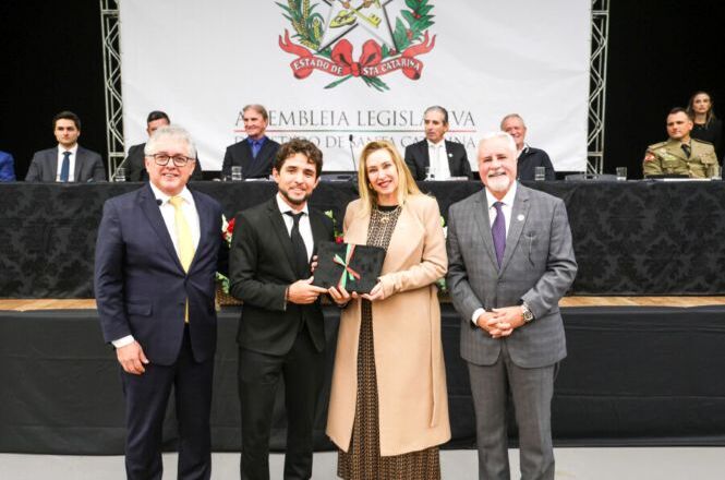 Foto horizontal: autoridades e homenageados posam com troféu durante cerimônia na Assembleia Legislativa, mesa de autoridades ao fundo, ambiente institucional, clima solene.