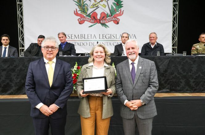 Foto horizontal: autoridade entrega certificado a homenageada durante cerimônia na Assembleia Legislativa, mesa de autoridades ao fundo, ambiente institucional, clima solene.
