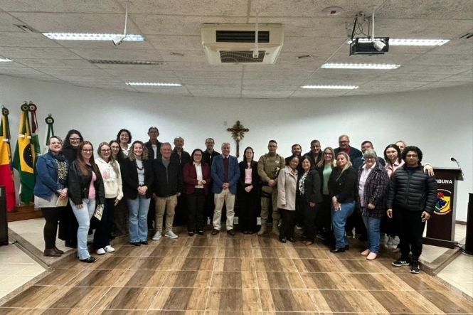 Foto horizontal: grupo de homens e mulheres posa no plenário da Câmara de Vereadores, com bandeiras ao fundo, após evento alusivo ao Agosto Lilás.