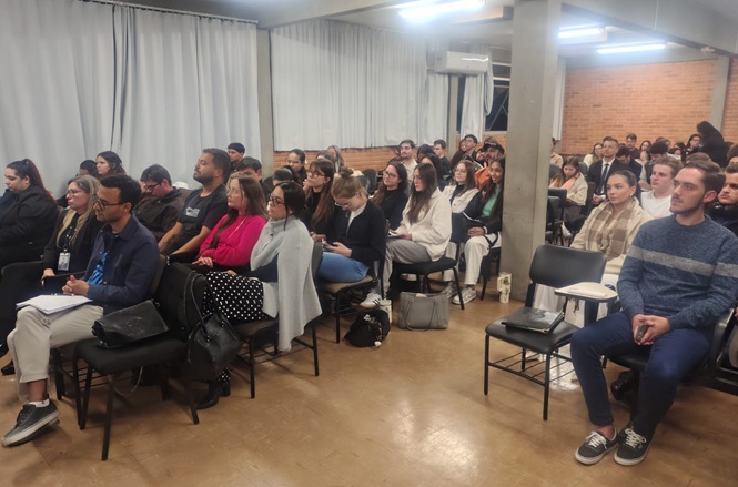 Foto horizontal: público acompanha apresentação em sala, participantes sentados e atentos, clima de aprendizado e troca de conhecimento.